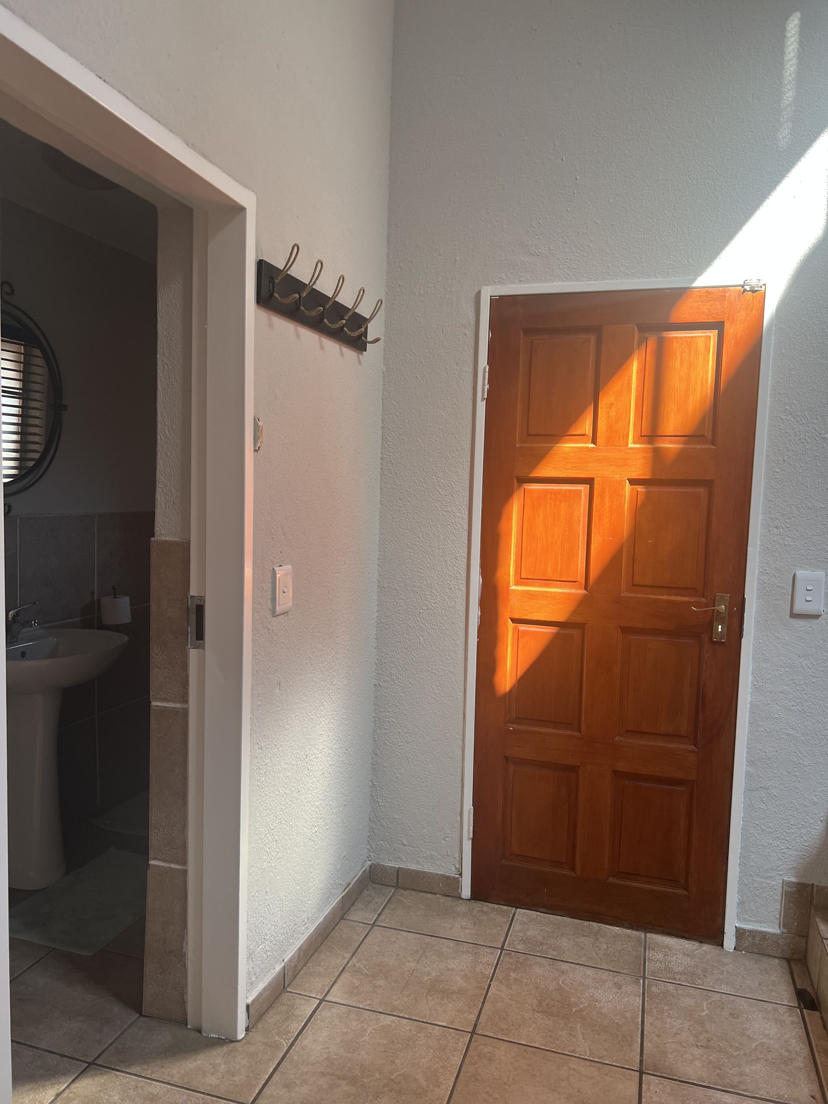 A hallway with a wooden door, a white wall, a small shelf with hooks, and a tiled floor.