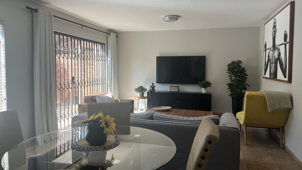A living room with a glass table, sofa, and a TV mounted on the wall. - Roodepoort Smart Home 1 (Roodepoort)