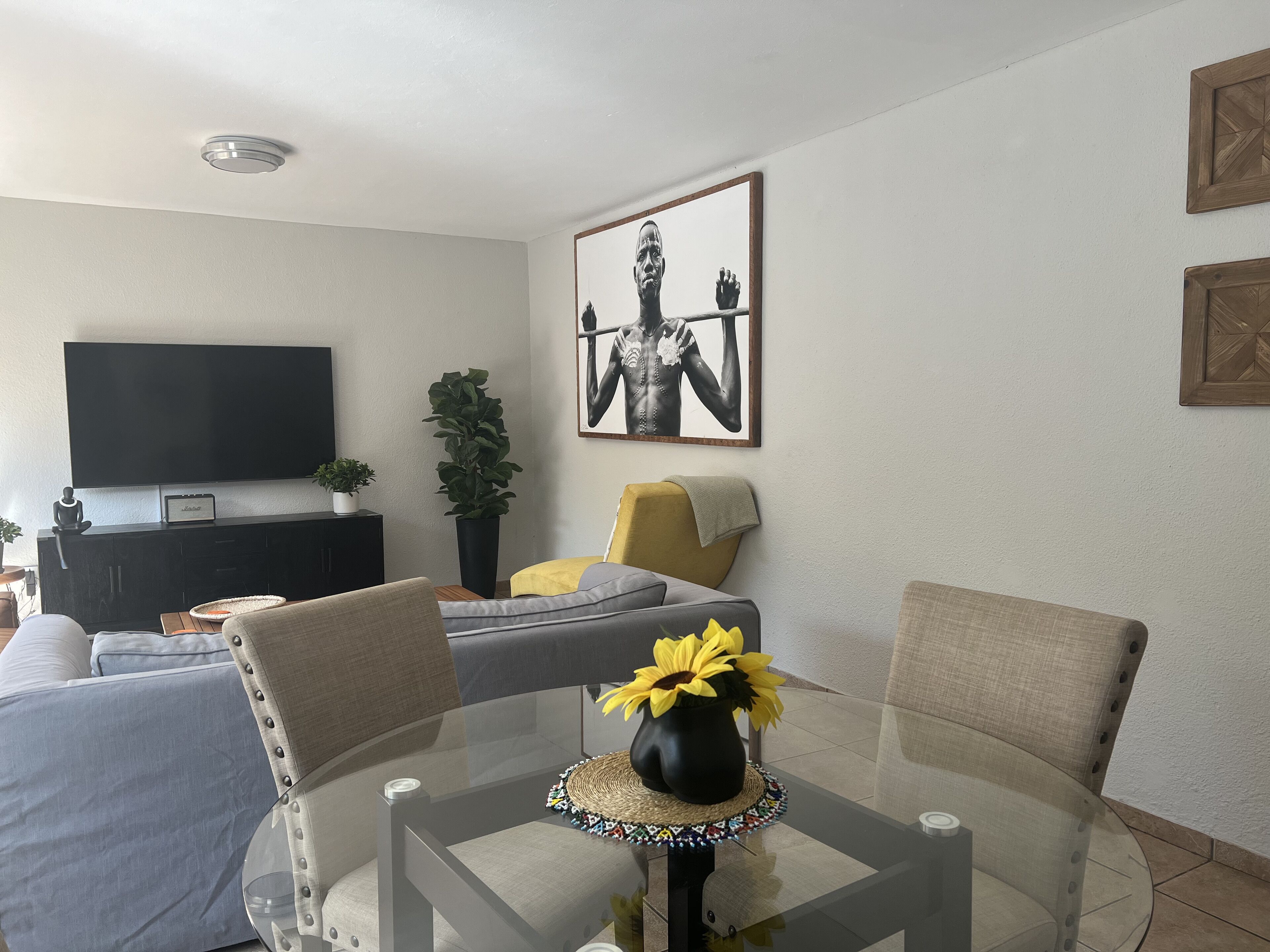 A modern living room with a flat-screen TV, a sofa, a dining table with chairs, and a large framed artwork on the wall.