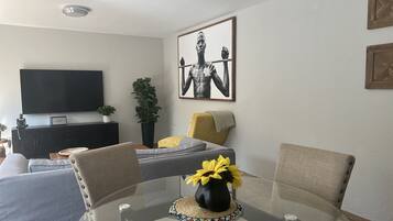 A modern living room with a flat-screen TV, a sofa, a dining table with chairs, and a large framed artwork on the wall.
