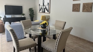 A dining area with a glass table surrounded by four upholstered chairs, a black cabinet with a flat-screen TV, and a potted plant. - Roodepoort Smart Home 1 (Roodepoort)