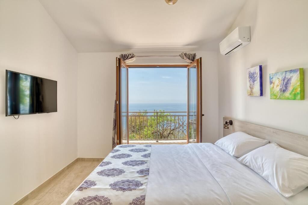 A bedroom with a large bed, a TV mounted on the wall, and a window with a view of the sea.