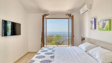 A bedroom with a large bed, a TV mounted on the wall, and a window with a view of the sea.