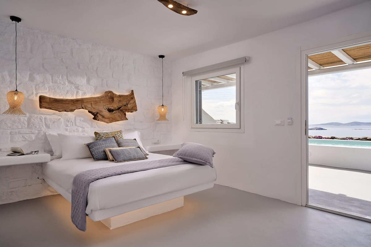 A bedroom with a bed, white walls, a wooden headboard, a window with a view of the sea, and pendant lights.