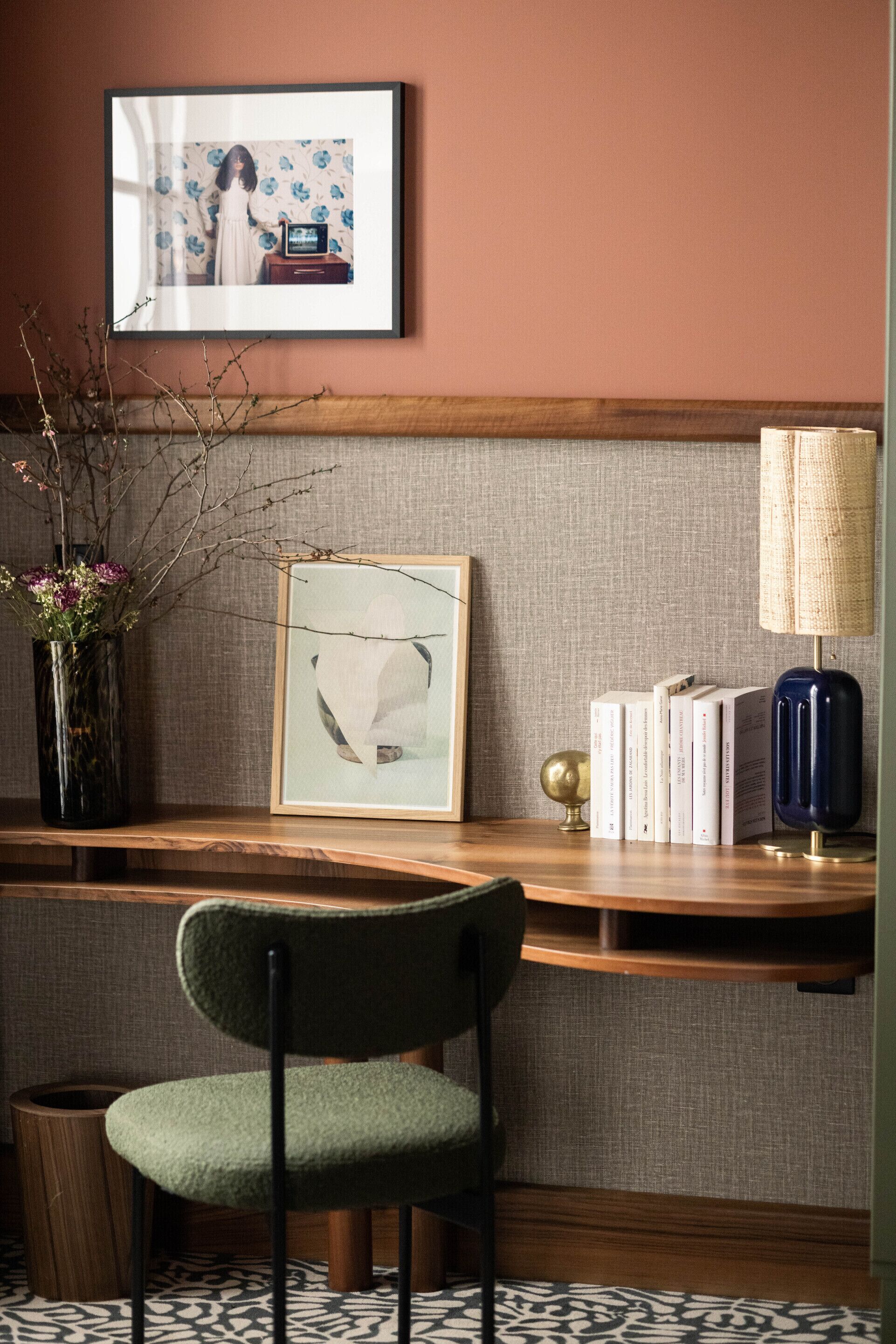 A hotel room with a wooden desk, a framed picture, a vase with flowers, a lamp, and a chair.