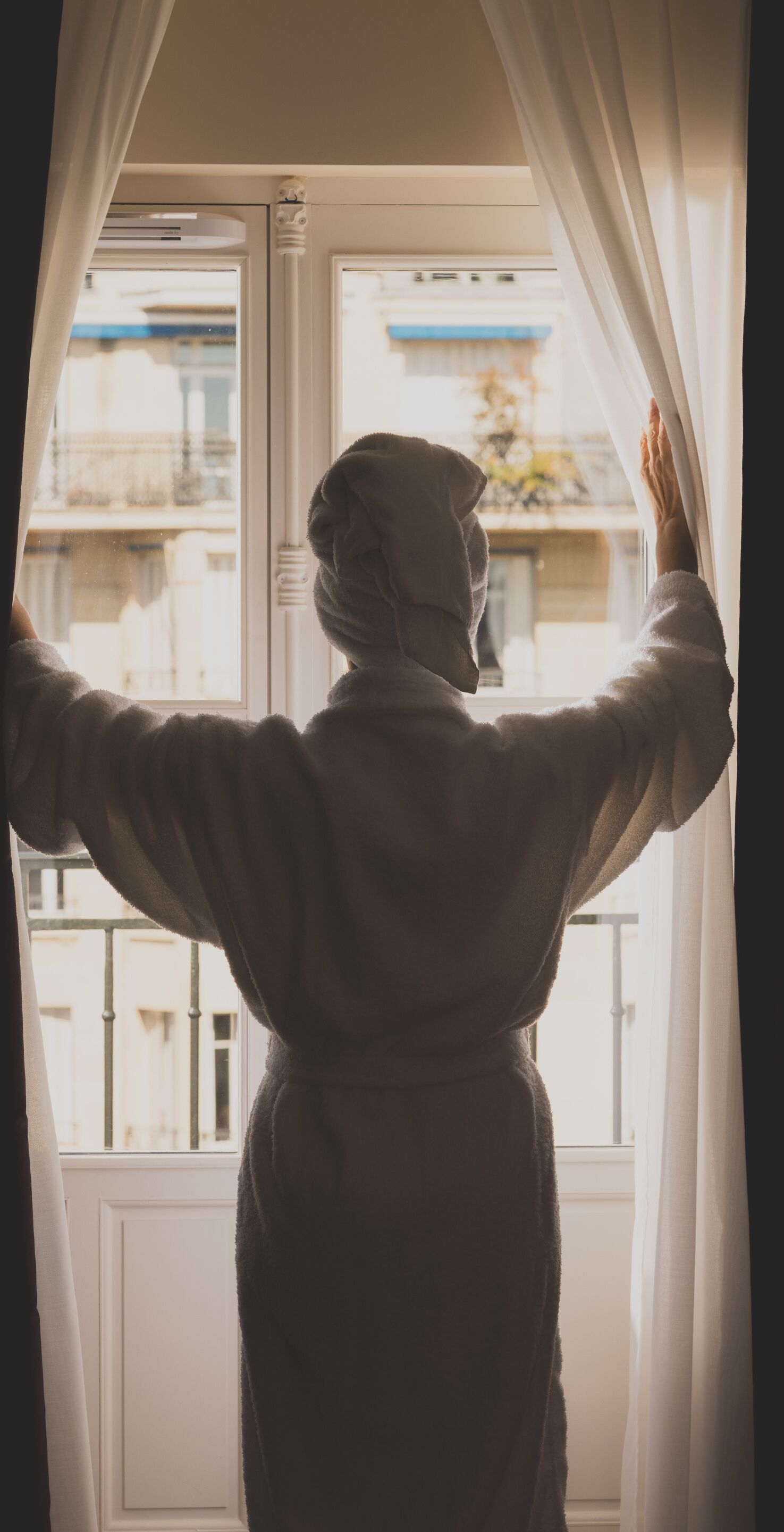 Una persona con bata de baño de pie junto a una ventana con cortinas, mirando al exterior.