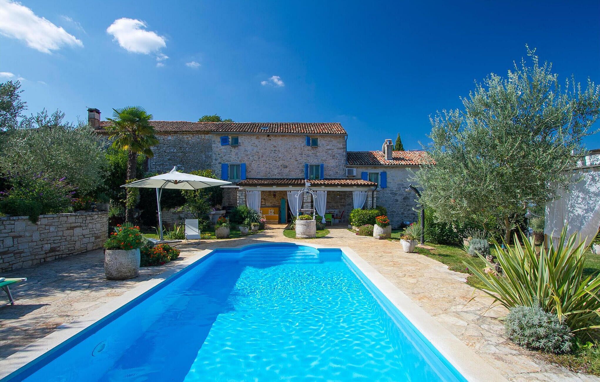 Una villa de piedra con piscina, rodeada de exuberante vegetación y un cielo azul claro.