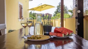 A table set with wine glasses, a pitcher, olives, cheese, and watermelon slices, with a yellow umbrella and lavender plants in the background.