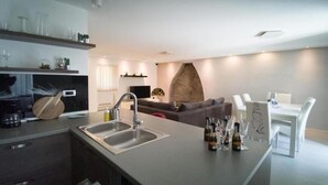 A modern kitchen with a large island, a dining area with a white table and chairs, and a living room with a sofa and a stone fireplace.
