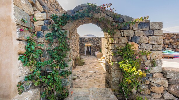 Un arco de piedra cubierto de hiedra, que conduce a un sendero de piedra y una vista de la montaña al fondo.