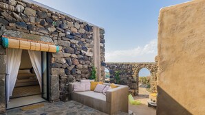 Una habitación con paredes de piedra, una zona de estar con cojines, una ventana con cortinas translúcidas y una vista a través de un arco.