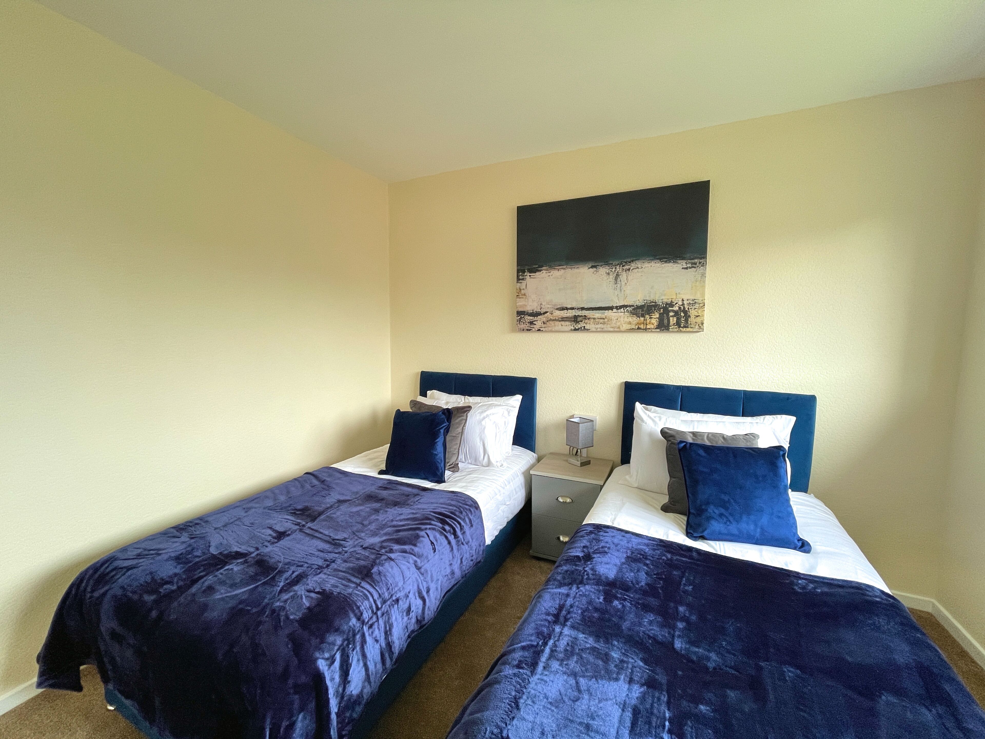 A room with two beds, each with a blue headboard and white bedding. There is a painting on the wall above the beds.