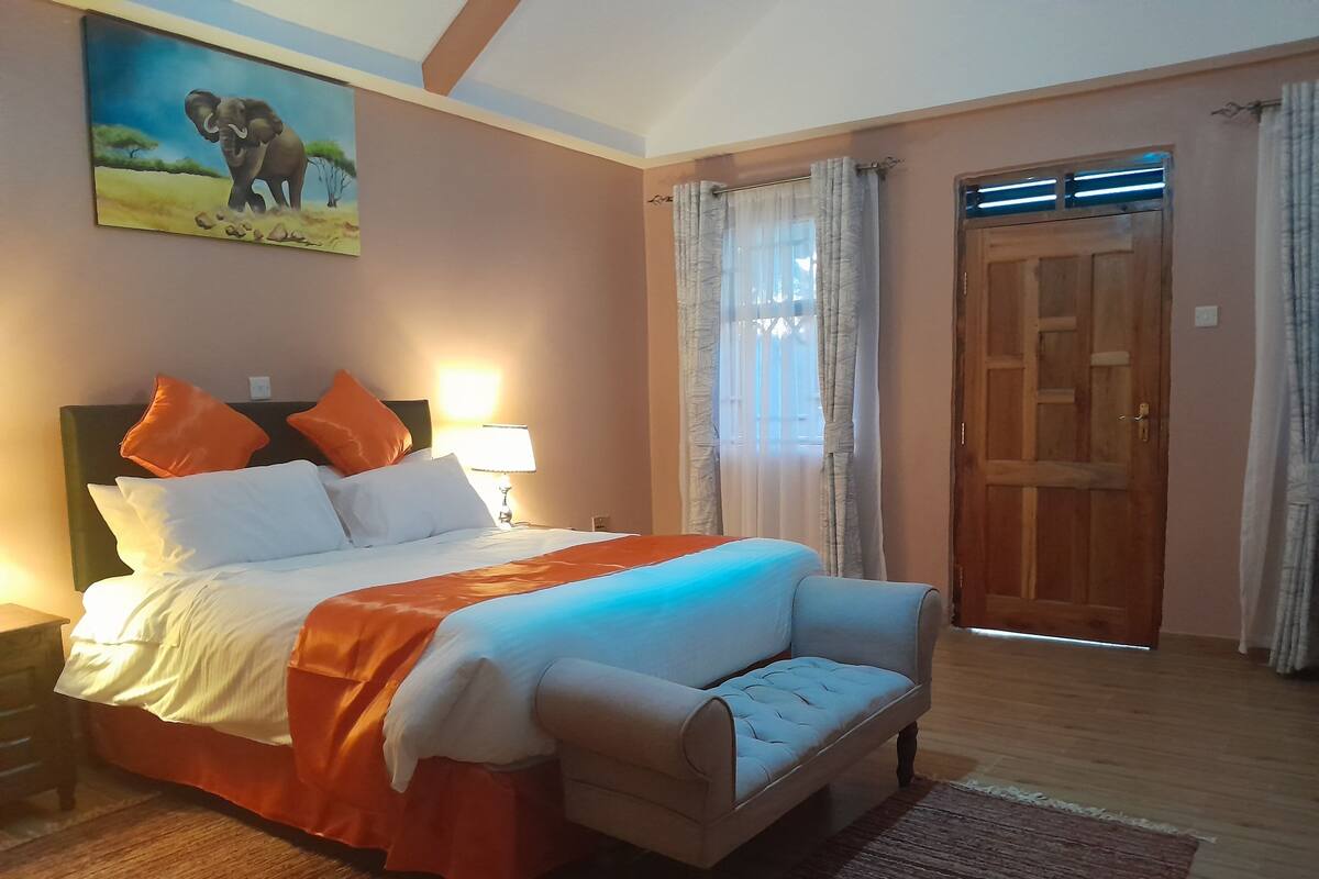 Un dormitorio con cama, almohadas, una mesita de noche, lámpara, un cuadro, ventana con cortinas y una puerta de madera.