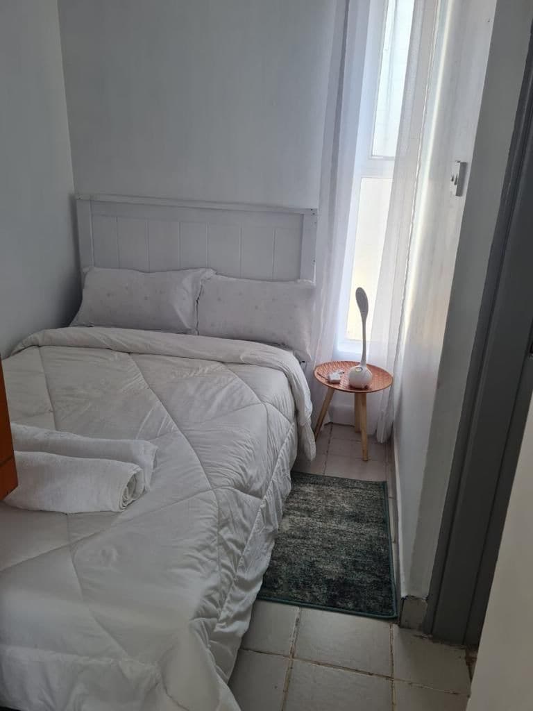 Un dormitorio con cama, mesita de noche, una pequeña mesa con silla y ventana con cortinas.