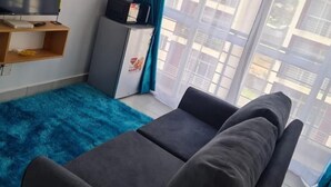 Un saló amb un sofà, televisió, nevera petita i una finestra amb cortines.