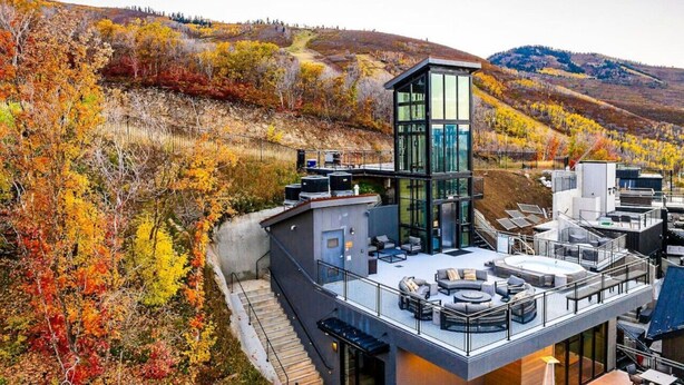 Un bâtiment moderne doté d’une façade en verre, d’un espace extérieur aménagé pour s’asseoir et offrant une vue sur une colline boisée.