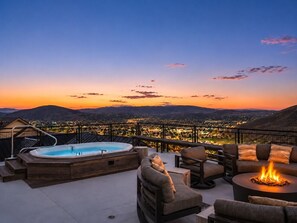 Terraza en la azotea con jacuzzi, mobiliario exterior y una chimenea, con vistas a un paisaje urbano al atardecer.