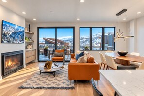 A modern living room with a fireplace, a sofa, a dining area, and large windows offering a mountain view. - Kings Crown Ski-In/Out•Private Hot Tub•Walk to Main (Park City)