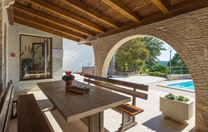 Um pátio ao ar livre com mesa e bancos de madeira, arcos de pedra, piscina e vista para áreas verdes e colinas.