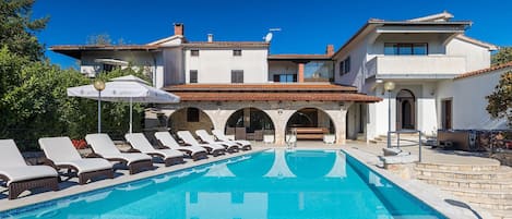 Une grande piscine bordée de chaises longues, un bâtiment orné d’arches et un ciel d’un bleu limpide.