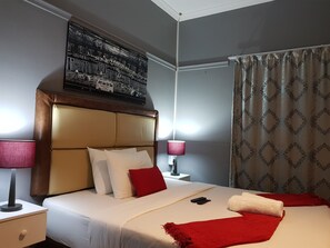 A hotel room with a large bed, two bedside tables, a nightstand, a red throw blanket, a white towel, and a painting on the wall. - Kingsworth Inn - Port Elizabeth (Gqeberha (Port Elizabeth))