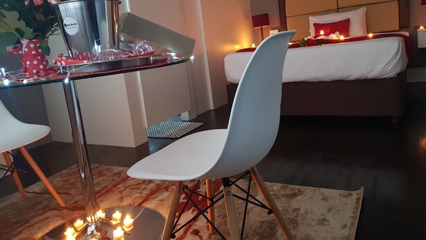 A modern bedroom with a bed, bedside table, and a chair. There is a small table with a bottle and glasses, and a vase with flowers. - Kingsworth Inn - Port Elizabeth (Gqeberha (Port Elizabeth))