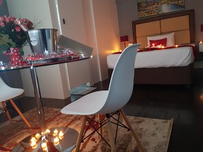 Une chambre à coucher moderne avec un lit, une table de chevet et une chaise. On y trouve une petite table avec une bouteille et des verres, ainsi qu’un vase rempli de fleurs.