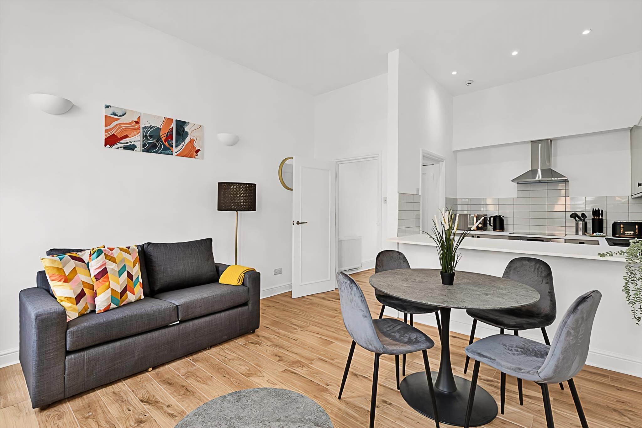 A modern living room with a grey sofa, colorful cushions, a round dining table with grey chairs, and a small side table with a plant.