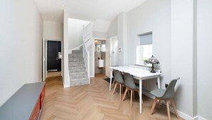 Un intérieur moderne avec un escalier menant à un deuxième étage, un coin repas avec une table blanche et des chaises grises, et une fenêtre agrémentée d’une plante.