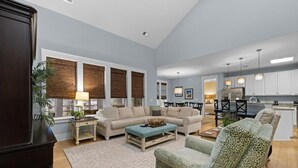 Amplia sala de estar con sofá, sillón y mesa de centro, una cocina al fondo y ventanas grandes con persianas.