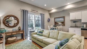 A cozy living room with a sofa, a coffee table, and a dining area with chairs. There is a kitchen with white cabinets and a window with curtains.