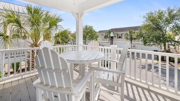 A covered outdoor seating area with white chairs and a table, surrounded by a white railing, overlooking a residential street with houses and palm trees. - Unwind in Style: Luxury 2 Bdrm Townhome Sleeps 8 (North Myrtle Beach)