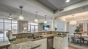 Una cocina moderna con encimeras de granito, armarios blancos y lavavajillas integrado. Se observan lámparas colgantes y un candelabro.