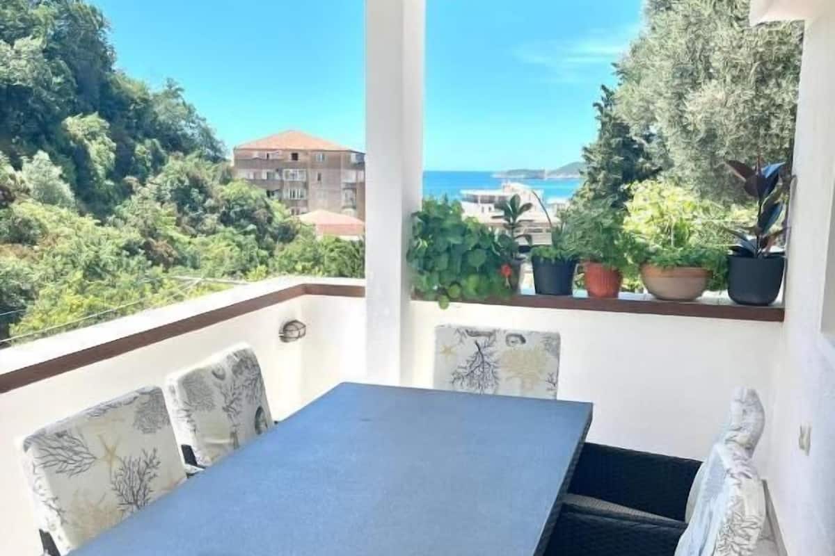 Ein Balkon mit Tisch und Stühlen, Blumentöpfen und Blick auf das Meer und Gebäude.