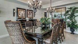 A dining room with a glass-top table, wicker chairs, a chandelier, and framed artwork on the walls. - Seaside Elegance: Oceanfront 5BR/5BA, Sleeps 14 (North Myrtle Beach)