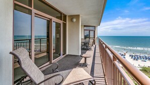 A balcony with wicker chairs and tables, overlooking a beach with umbrellas and people. - Seaside Elegance: Oceanfront 5BR/5BA, Sleeps 14 (North Myrtle Beach)
