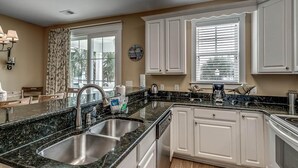 A kitchen with a granite countertop, stainless steel sink, white cabinetry, and a clock on the wall.