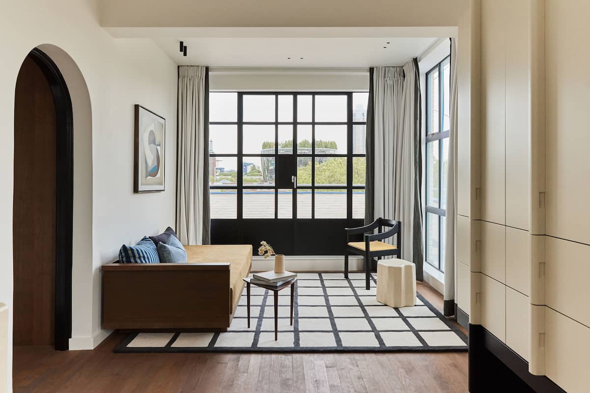 A modern living room with a sofa, a small table, and a chair. There is a large window with curtains, a framed picture on the wall, and a view of buildings and trees outside.