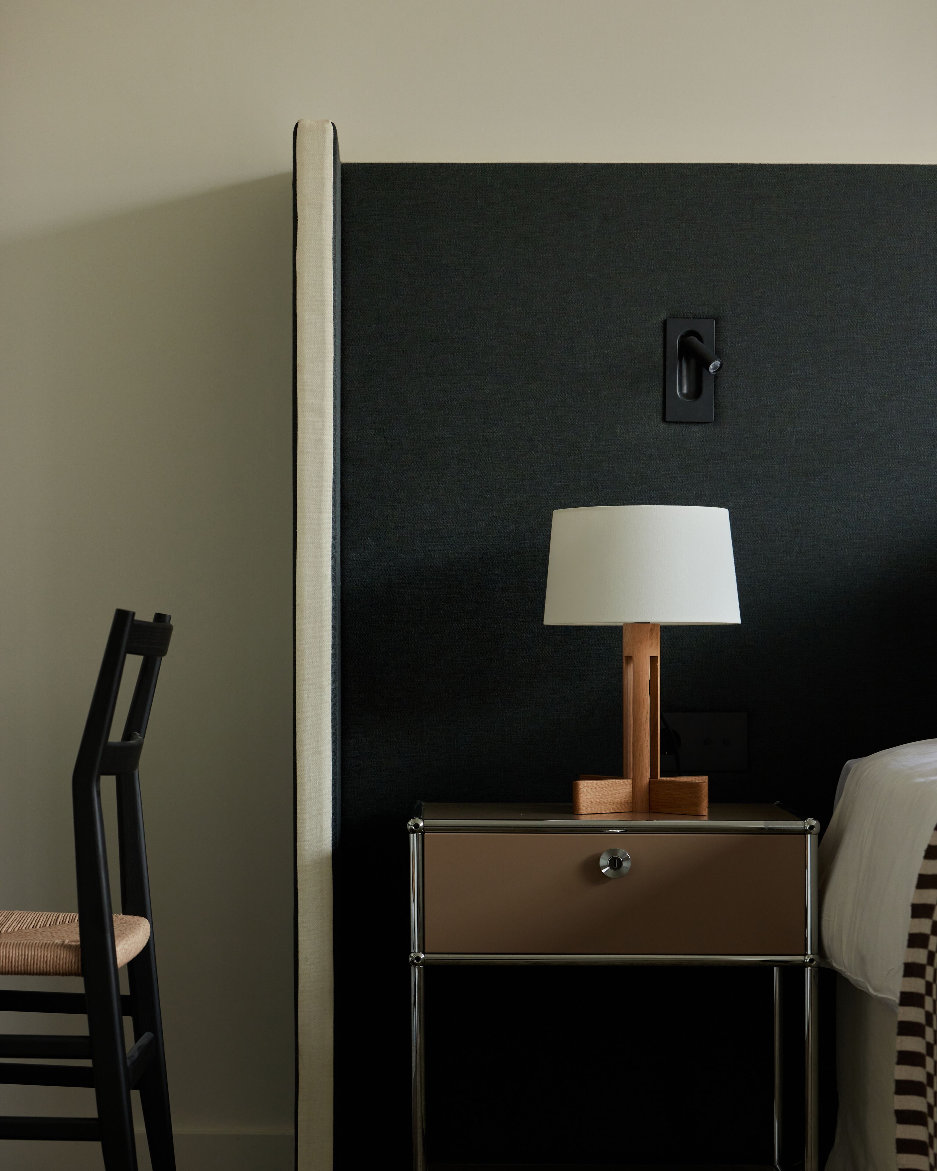 A hotel room with a dark wall, a wooden nightstand with a lamp, a chair, and a bed with a checkered bedspread.