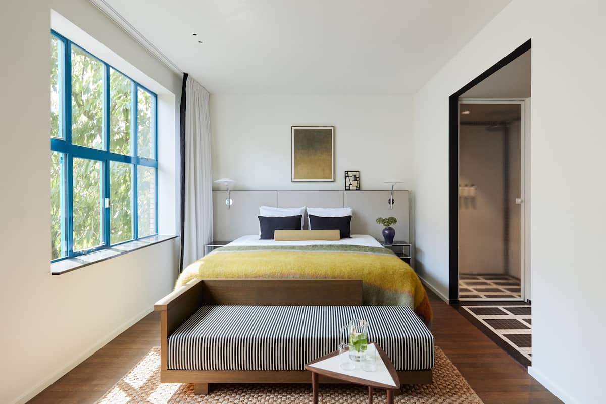 A modern hotel room with a large bed, a bench, a small table, and a view of the outdoors through a large window.
