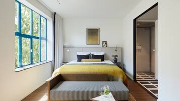 Una habitación de hotel moderna con una cama grande, un banco, una mesita pequeña y vistas al exterior a través de un amplio ventanal.