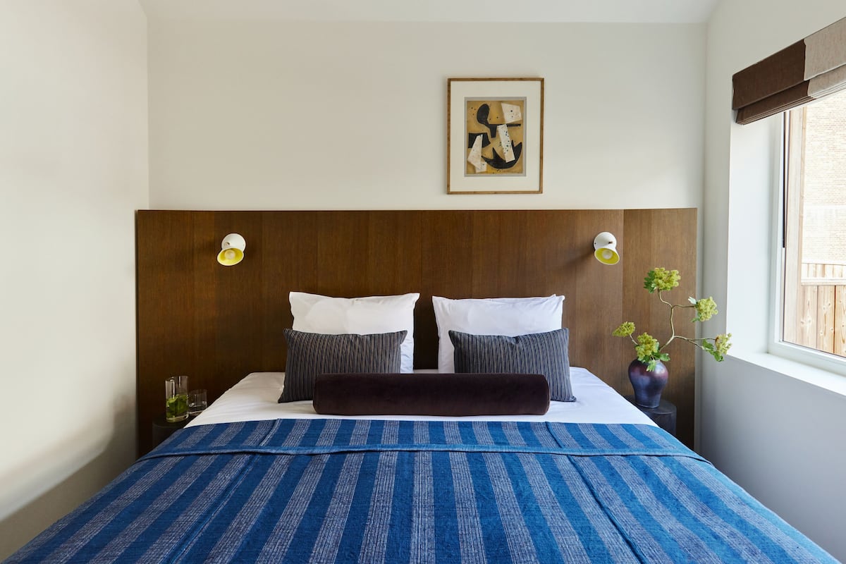 A hotel room with a large bed, a wooden headboard, two bedside lamps, a framed artwork above the bed, and a window with a view.