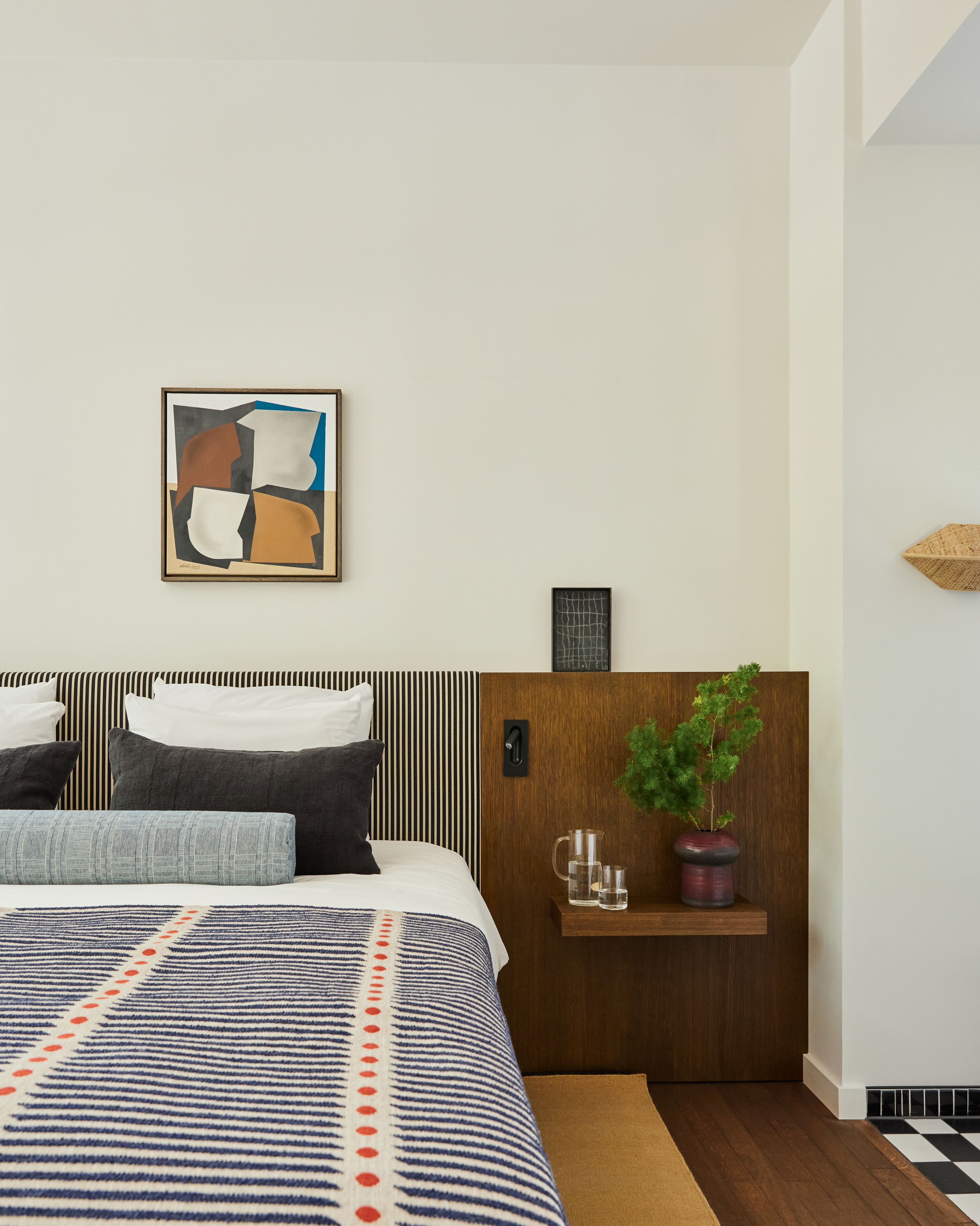 Een moderne hotelkamer met een gestreept hoofdeinde, een groot abstract schilderij, een houten plank met een potplant en een geruit vloertje.