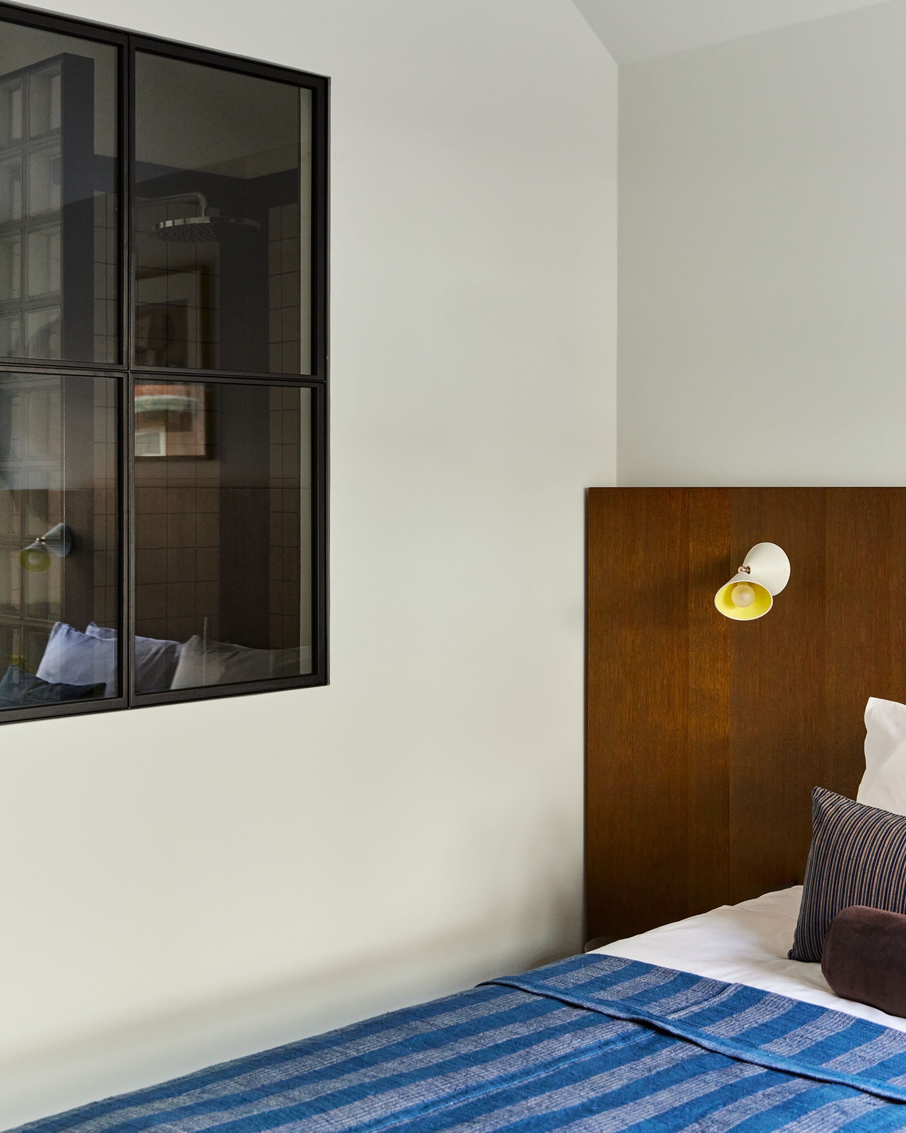 Habitación de hotel con un ventanal grande, cabecero de madera, lámpara de noche y cama con manta azul a rayas y una almohada.