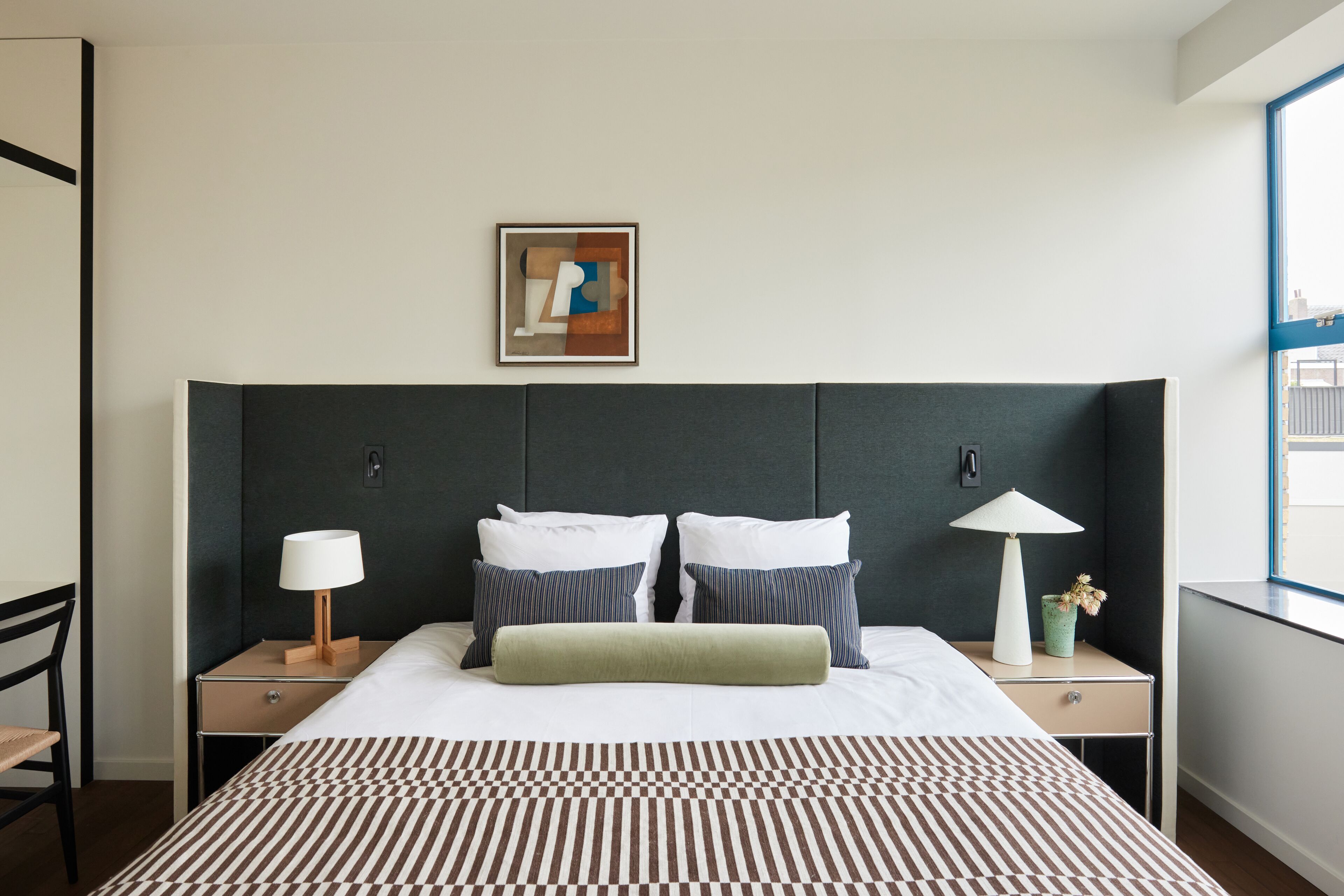 Une chambre d’hôtel avec un grand lit, deux tables de chevet, une commode, une lampe, un tableau encadré au mur et une fenêtre donnant sur un beau paysage.