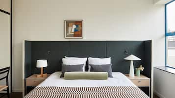 Une chambre d’hôtel avec un grand lit, deux tables de chevet, une commode, une lampe, un tableau encadré au mur et une fenêtre donnant sur un beau paysage.
