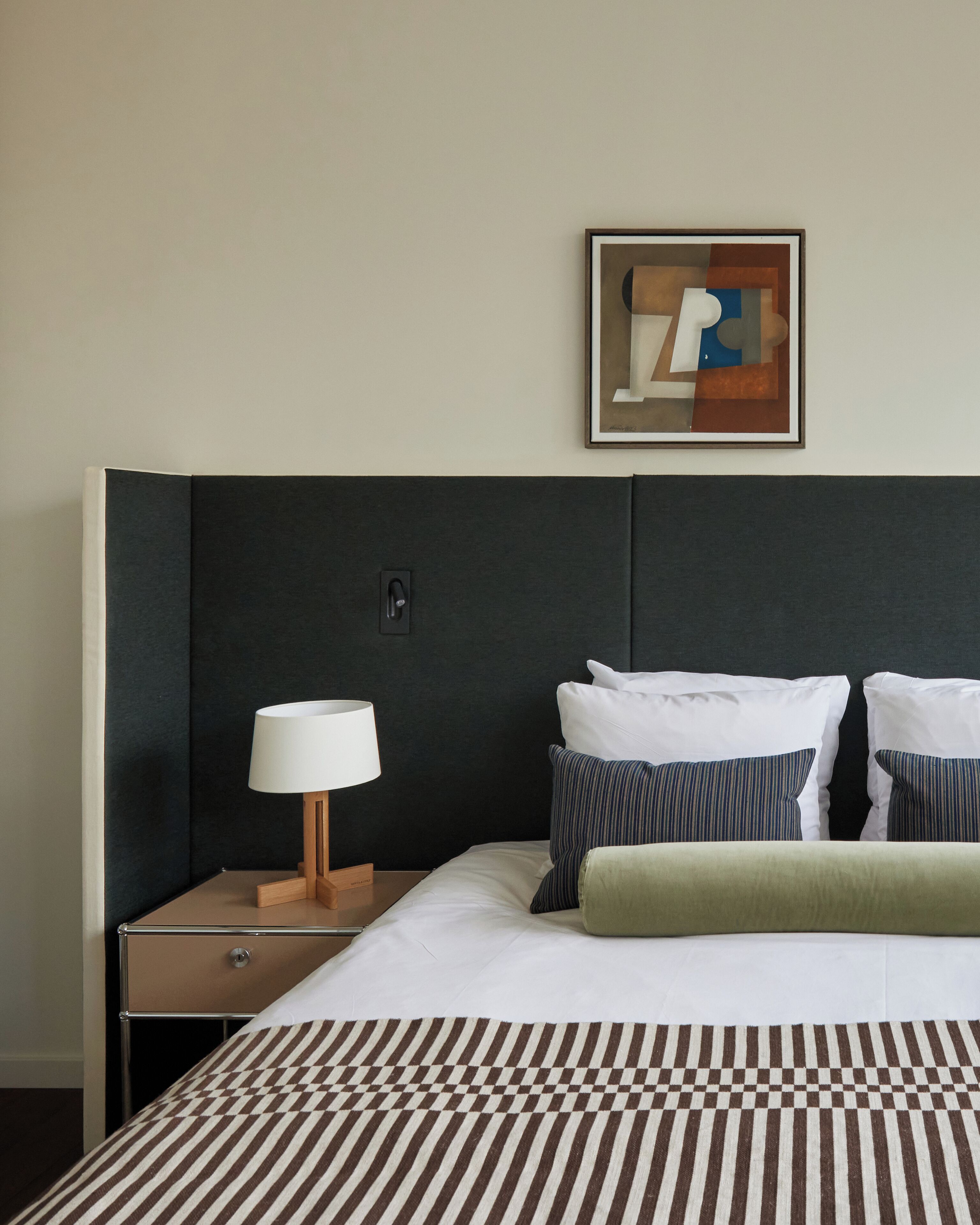 Uma cama bem arrumada com roupa de cama listrada, uma luminária de cabeceira e um quadro abstrato na parede.