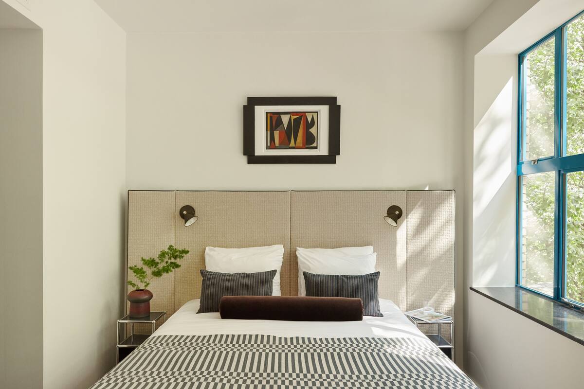 A modern hotel room with a large bed, a framed artwork above the bed, a bedside table with a plant, and a window with a view of trees.