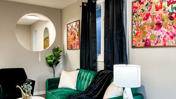 A modern living room with a green sofa, a round mirror, a white lamp, and a floral painting. - Charming 2BR Home | Sleeps 5| Full Stock Kitchen (Drumheller)