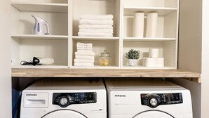 A laundry area with two Samsung VRT washers, shelves above holding towels, toilet paper, and a small plant. - Charming 2BR Home | Sleeps 5| Full Stock Kitchen (Drumheller)